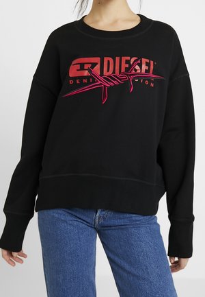 Czarna bluza z długim rękawem z czerwonym logo Diesel i grafiką drutu kolczastego, zestawiona z niebieskimi dżinsami na stojącej osobie.