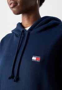 Primo piano di una persona che indossa una felpa con cappuccio blu scuro con la toppa del logo "Tommy Jeans" sul petto e i cordini del cappuccio appesi.