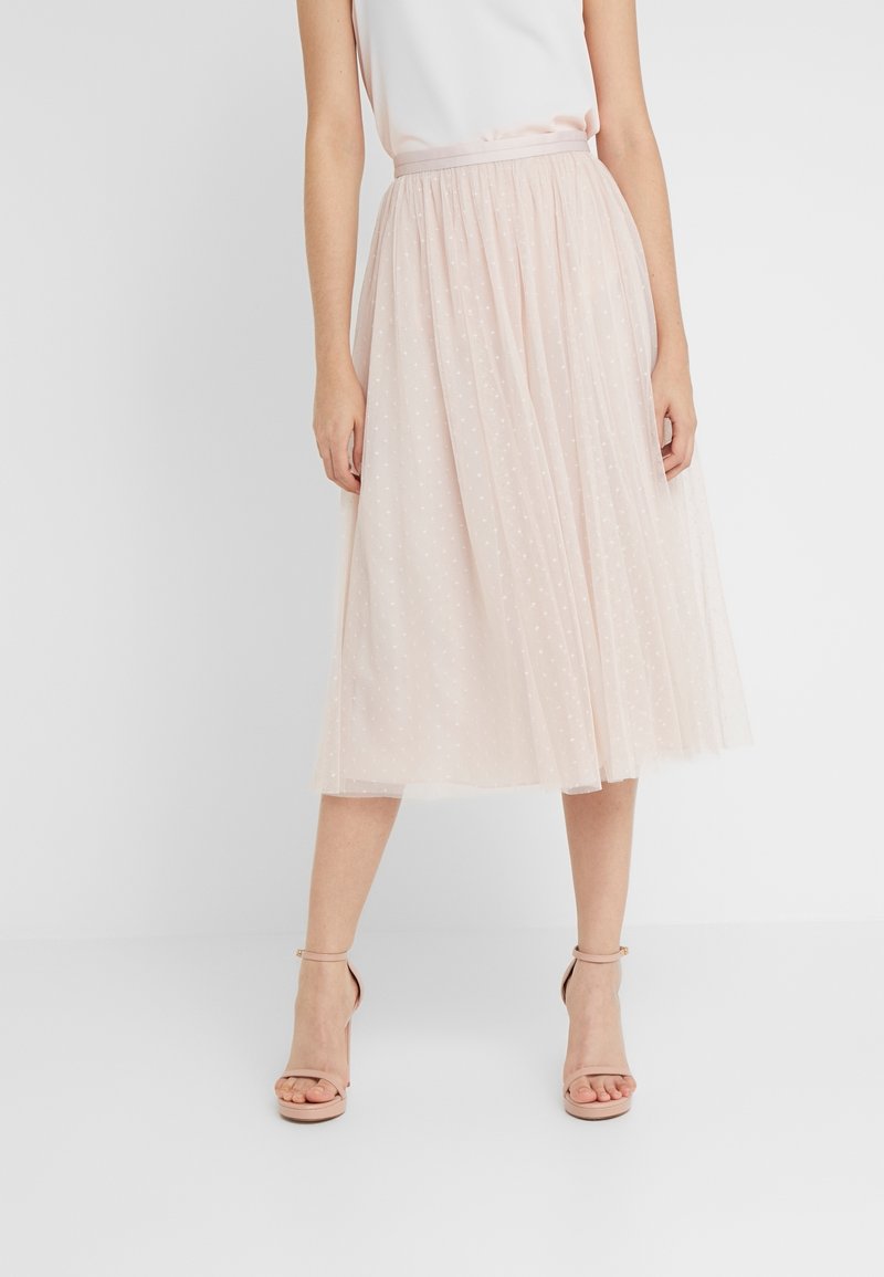 Femme portant une jupe midi en tulle rose pâle avec des pois subtils, associée à des talons hauts nude à bride cheville sur fond blanc.