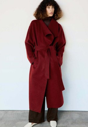 Un mannequin se tient devant un mur uni, vêtu d’un long manteau bordeaux ceinturé, d’un pantalon sombre et de chaussures blanches.