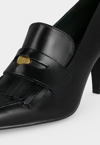 Μαύρο δερμάτινο loafer με τακούνι, μυτερή μύτη, διακοσμητικό με κρόσσια και χρυσό στρογγυλό έμβλημα στο λουρί.