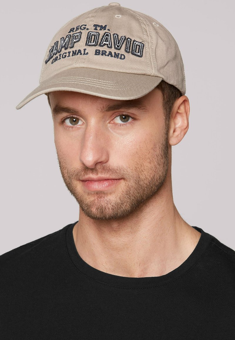 Cappellino da baseball in cotone beige con ricamo "CAMP DAVID" in blu navy, visiera curva e sei fori di ventilazione. Abbinato a una t-shirt nera.