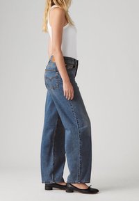 Femme debout en jean bleu à jambes larges, haut blanc sans manches et chaussures noires à talons bas sur un fond clair uni.
