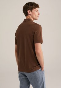 Camisa polo marrón con un cuello clásico y mangas cortas, combinada con pantalones cortos grises. El material parece suave con una textura lisa.