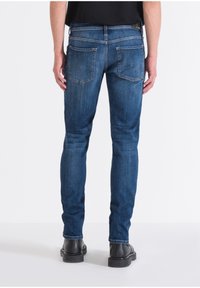 Jeans de mezclilla azul con corte slim, que cuentan con dos bolsillos traseros y costuras de contraste. Llevados con zapatos negros y una camiseta negra de manga corta.