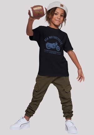 Black t-shirt with blue graphic of a motorcycle, green cargo pants, and white sneakers. Child holding a football, wearing a light-colored beanie.