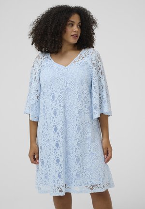 Kobieta o kręconych włosach, w jasnoniebieskiej koronkowej sukience w kwiaty z rozkloszowanymi rękawami do łokcia, stojąca na jednolitym tle.