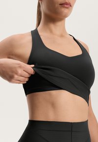 Čierny športový podprsenka s výstrihom do V, hladká tkanina a bezšvové detaily. Modelka si upravuje tkaninu, aby odhalila spodnú vrstvu.
