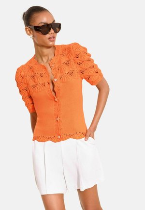 Femme portant de grandes lunettes de soleil foncées, un haut orange en maille à manches courtes avec boutons et détails en dentelle, et un short blanc taille haute.