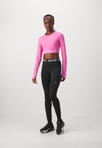 Nike Pro crop top i lys rosa, kombinert med svarte high-waisted leggings med Nike-logo. Modellen har på seg svarte treningssko.