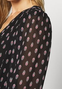 Zwarte doorschijnende stof met een herhalend bloemenpatroon in wit en rood. De blouse heeft zachte plooien op de schouders voor extra textuur.