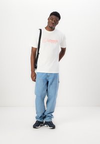 White cotton t-shirt with red text, loose blue denim cargo pants, and black athletic shoes. The model stands against a plain background.