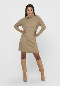 Beige strikket turtleneck kjole med droppe skuldre, ribbestruktur og lengde til midten av lårene, kombinert med brune knehøye støvler.