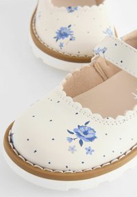 Weiße Lederschuhe mit blauem Blumenmuster, gewelltem Rand und beige Gummisohle. Sie verfügen über einen Riemen über dem Spann für einen sicheren Halt.