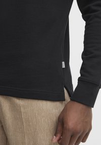 Black long-sleeve shirt made of cotton, featuring a side split hem and stitched detailing, paired with beige textured trousers.