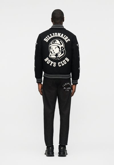 Άνδρας που φοράει μαύρο μπουφάν Billionaire Boys Club με λογότυπο αστροναύτη και μαύρο τζιν με λευκό κείμενο λογότυπου στην πίσω τσέπη, στέκεται με την πλάτη προς τα εμπρός.