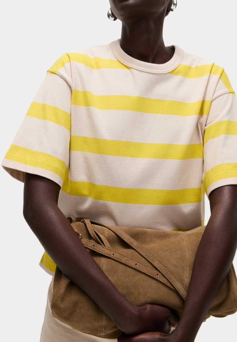 Béžové bavlněné tričko se širokými žlutými pruhy, krátkými rukávy a kulatým výstřihem; drženo hnědou semišovou kabelkou.