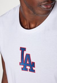 Bílý bavlněný tričko s výrazným vyšitým logem "LA" v červené a modré barvě na hrudi. Krátké rukávy a klasický kulatý výstřih.