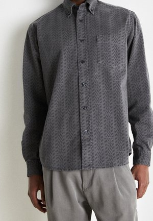 Długa koszula na guziki w ciemnoszarym kolorze z subtelnym wzorem liści i kieszenią na piersi, noszona z jasnoszarymi spodniami.