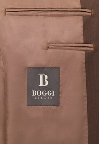 Brūns audums ar divām kabatām, ar pelēku etiķeti, uz kuras ir teksts "BOGGI MILANO" un stilizēts "B" logo. Gluda tekstūra.