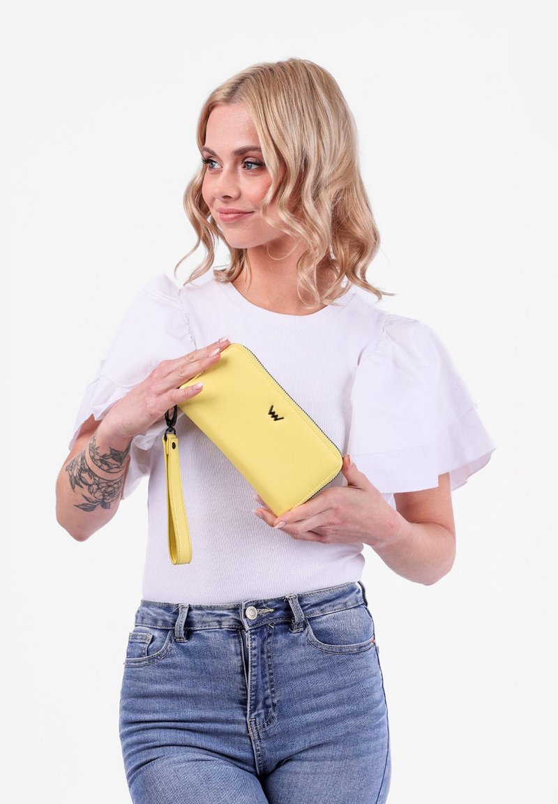 Yellow leather wallet with a smooth texture, zip closure, and wrist strap, featuring a subtle logo. Model holds it against a white background.