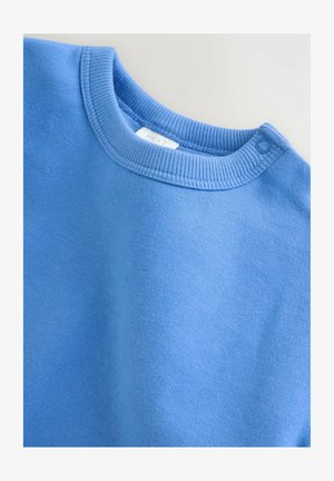 Svetlo modra bombažna majica s potiskanim okroglim izrezom in zapiranjem z zatiči na rami. Mehka tekstura z enakomerno barvo.