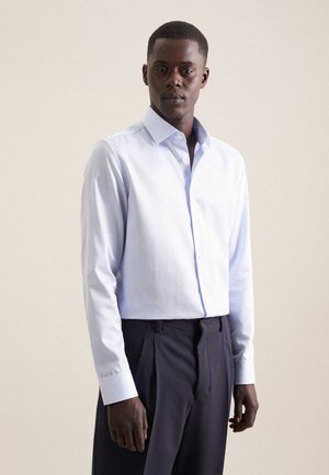 Homme portant une chemise à rayures bleu clair à manches longues et un pantalon plissé foncé, debout devant un fond clair uni.