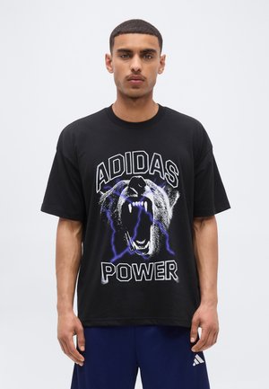 Mężczyzna w czarnej koszulce Adidas z grafiką ryczącego niedźwiedzia i niebieskich błyskawic, w połączeniu z niebieskimi spodniami Adidas.