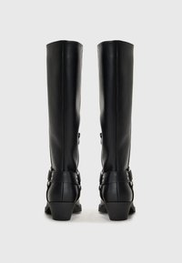 Botas negras hasta la rodilla hechas de material sintético suave. Presentan un tacón bajo y correas decorativas con botones metálicos a los lados.
