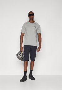 T-shirt gris à manches courtes avec un logo, associé à un short foncé. Tenant un casque gris, portant des lunettes de soleil. Des chaussures et des chaussettes sombres complètent la tenue.