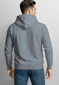 Grauer Kapuzenpullover mit langen Ärmeln, abgerundetem Saum und subtiler Textur, kombiniert mit blauen Jeans, die die Steppnähte der Gesäßtaschen betonen.