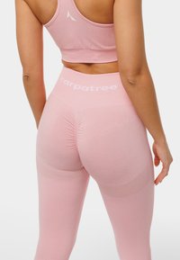 Leggings de sport roses et crop top assorti en matériau extensible, avec un détail froncé à l'arrière et des accents de logo blancs.