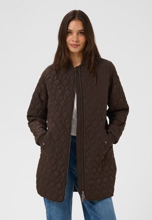 Femme portant une longue veste matelassée marron foncé avec poches avant et une fermeture éclair, associée à un jean bleu et un haut blanc.