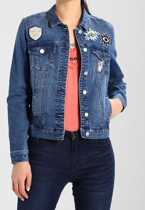 Femme portant une veste en jean bleue avec des patchs colorés sur un top corail et un jean foncé, debout avec une main dans la poche.