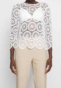 Blouse en dentelle blanche à manches longues, ornée de découpes circulaires florales et d'un ourlet festonné. Associée à un pantalon beige à rayures.