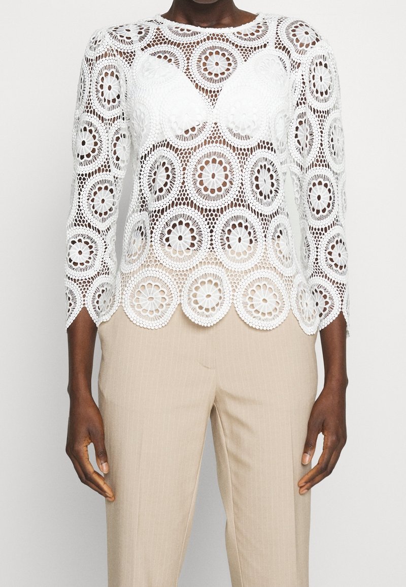 Blouse en dentelle blanche à manches longues, ornée de découpes circulaires florales et d'un ourlet festonné. Associée à un pantalon beige à rayures.