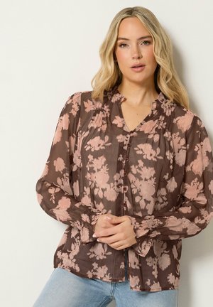 Kvinde med langt blondt hår iført en gennemsigtig brun bluse med lyserøde blomster og lyseblå jeans, står mod en ensfarvet lys baggrund.