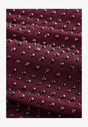 Bordó szövet texturált szövési mintázattal, fényes fekete és mélyvörös strasszokkal díszítve, amelyek szórtan helyezkednek el a felületen.