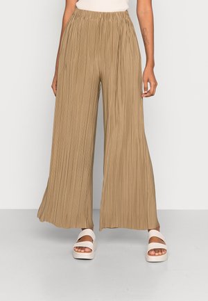 Beżowe, szerokie spodnie plisowane z elastycznym pasem, noszone z białymi sandałami na platformie z odkrytymi palcami na jasnym tle.
