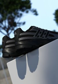 Čierne atletické topánky s vrchnou časťou z materiálu so sieťovinou, s šedými akcentmi a odolnou podrážkou. Topánky sú umiestnené na bielom podstavci.