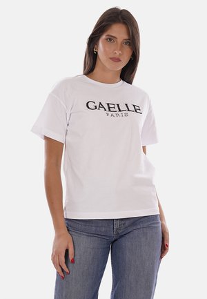 Femme aux cheveux châtain foncé portant un t-shirt blanc GAELLE PARIS et un jean bleu, debout avec une main détendue sur le côté.
