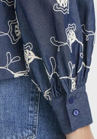 Blå denimbluse med ærme og hvid blomsterbroderi samt knappet manchet, hvilet nær lommen på blå denimjeans.