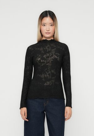 Kvinne iført en svart langermet blomstermønstret blondebluse og mørkeblå jeans, stående mot en enkel lys bakgrunn, vendt fremover.