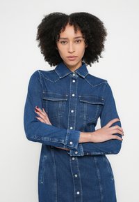 Tumesinises toonis teksariidest särk-kleit nööpidega ees, kahe rinnatasku ja pikade varrukatega, millel on nööbitavad mansetid.