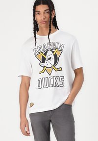 T-shirt bianco in cotone con logo "Anaheim Ducks", grafiche nere e gialle, vestibilità larga, maniche corte, colletto rotondo e jeans in denim grigi.