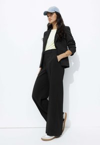 Blazer noir ajusté porté sur un t-shirt jaune clair, associé à un pantalon large noir et des baskets blanches avec une semelle en caoutchouc. Une casquette bleue complète le look.