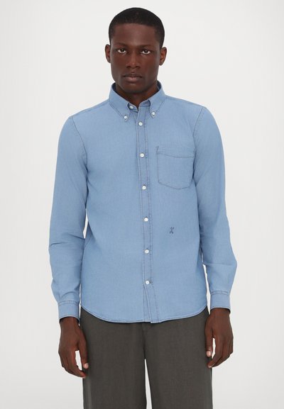 Light blue denim shirt with a button-down collar, long sleeves, and a chest pocket. Features visible stitching and a curved hemline.