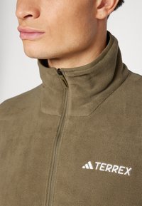Fleecejakke i olivengrøn med høj krave og frontlynlås. Har hvid "TERREX" logo og struktureret stof.