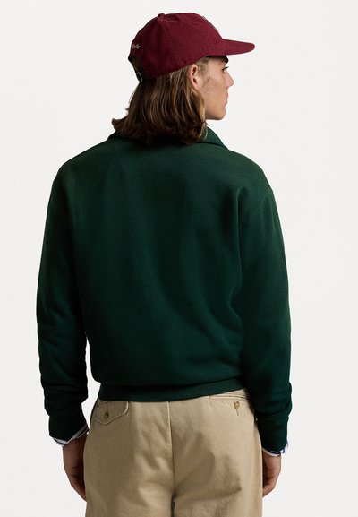 Felpa verde con maniche lunghe, polsini a coste, vestibilità rilassata, abbinata a pantaloni beige chiaro e un berretto bordeaux. Texture sottile visibile.