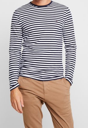 Long-sleeve navy and white striped t-shirt with a round neckline and a small logo on the chest, paired with beige pants.